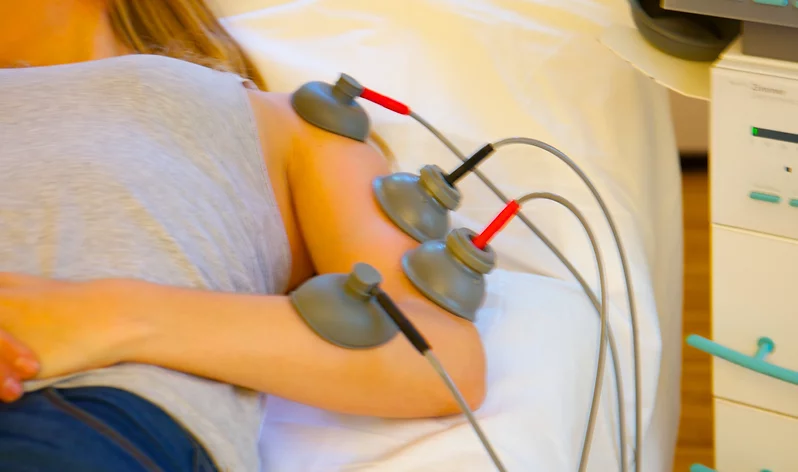 Durchführung einer Elektrotherapie. Elektroden sind am Arm einer auf einer LIege liegenden Frau befestigt.